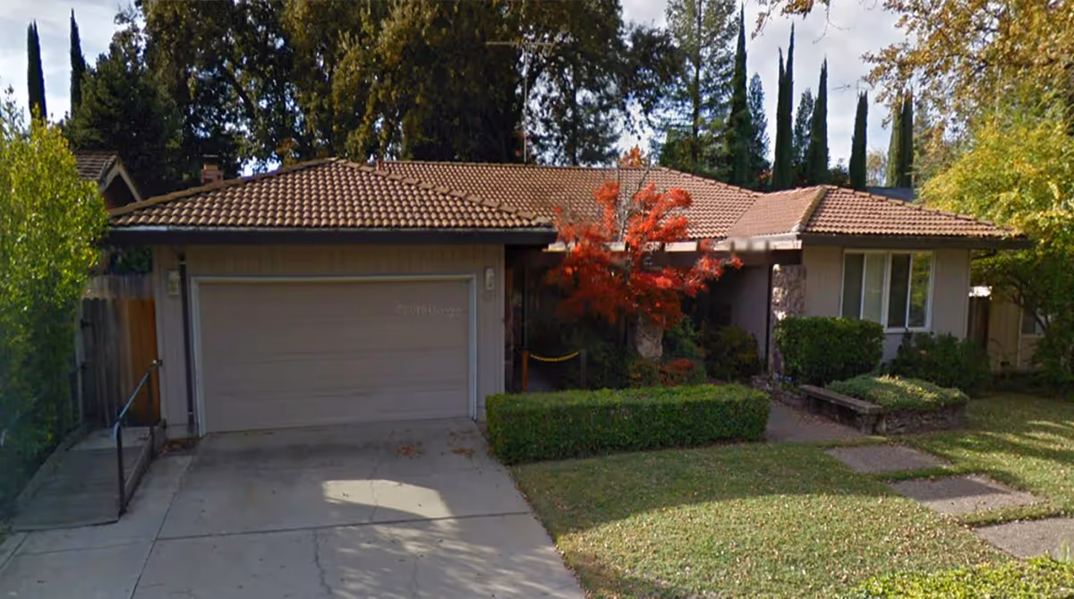 Front exterior view of a single-story house with a tiled roof, a two-car garage, a driveway, and a well-maintained lawn with bushes and a small tree with red leaves.