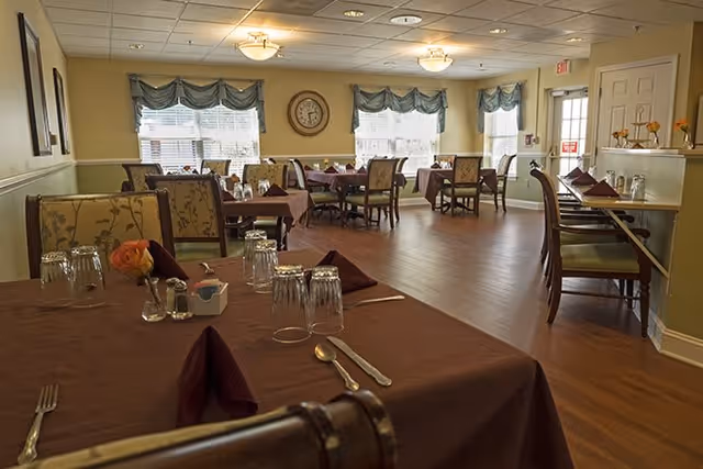 A dining room with several tables covered in brown tablecloths, each set with upside-down glasses, silverware, and folded napkins. The room has wooden flooring, large windows with valances, wall clock, and soft ceiling lights. Chairs with floral upholstery surround the tables.