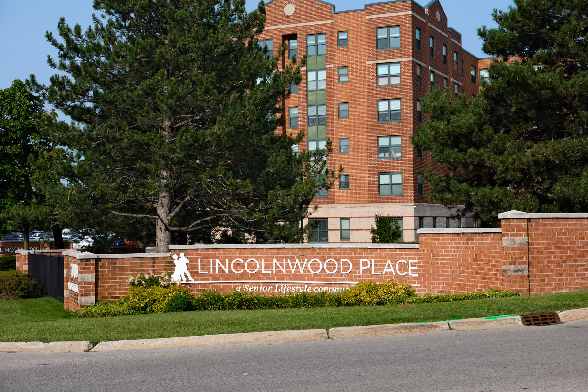 Exterior view of Lincolnwood Place senior living community showing a brick sign with the community name and a multi-story brick building in the background surrounded by trees and greenery.