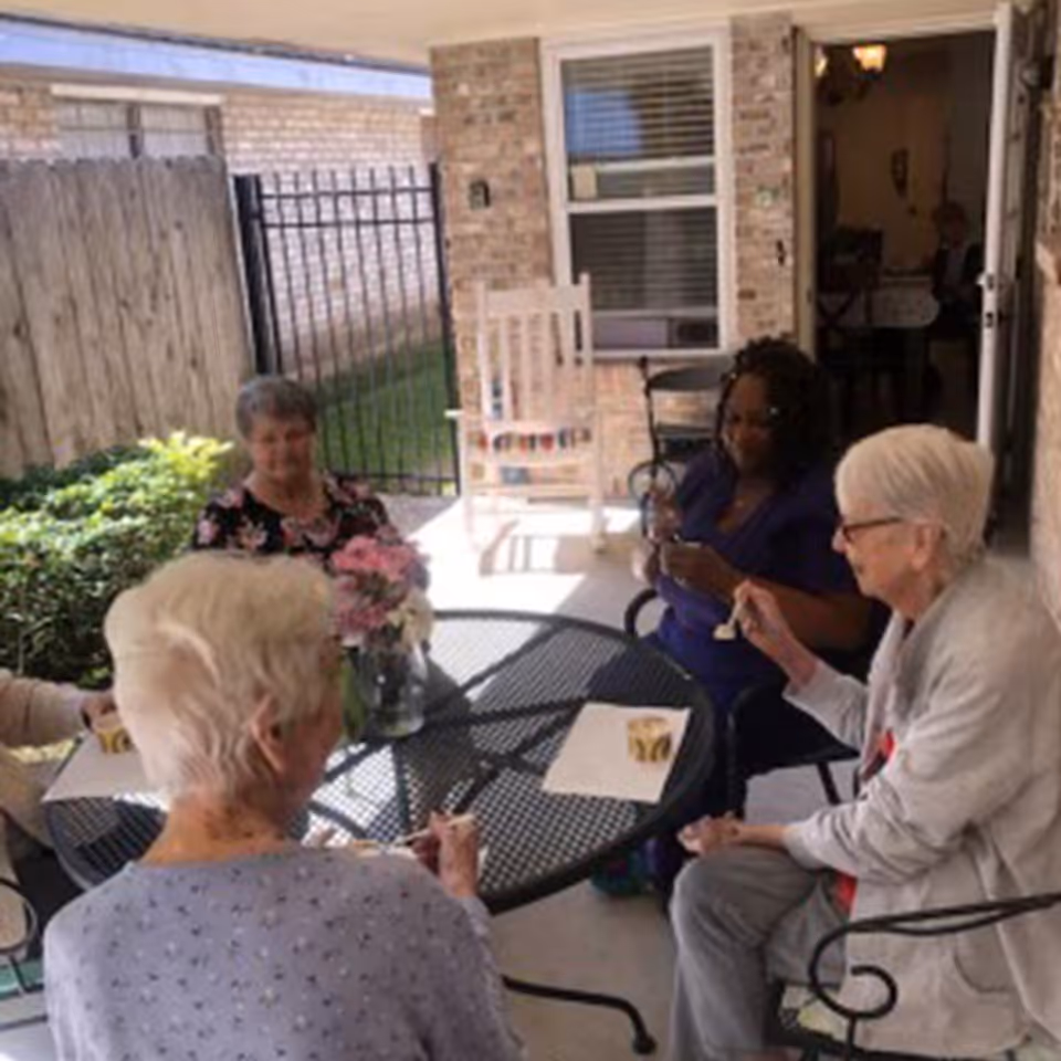 Four elderly women sitting around a round metal table on a covered patio, enjoying a snack together. There is a vase with pink flowers on the table. The patio is enclosed with a wooden fence and a black metal gate, and a brick wall with a window and an open door leading inside is visible in the background.