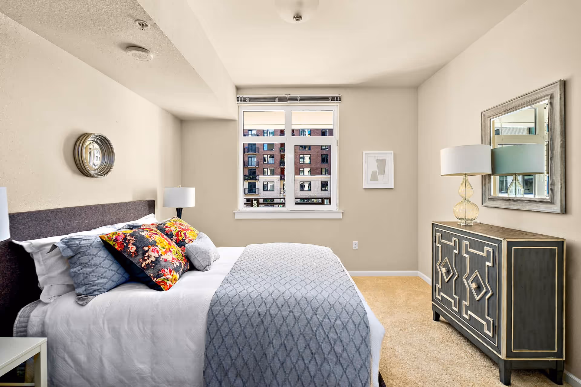 A bright bedroom with a neatly made bed featuring white sheets, a gray patterned blanket, and colorful floral pillows. There is a round decorative mirror on the wall above the bed, a window showing an exterior view of a building, and a dresser with a table lamp and a framed mirror above it.