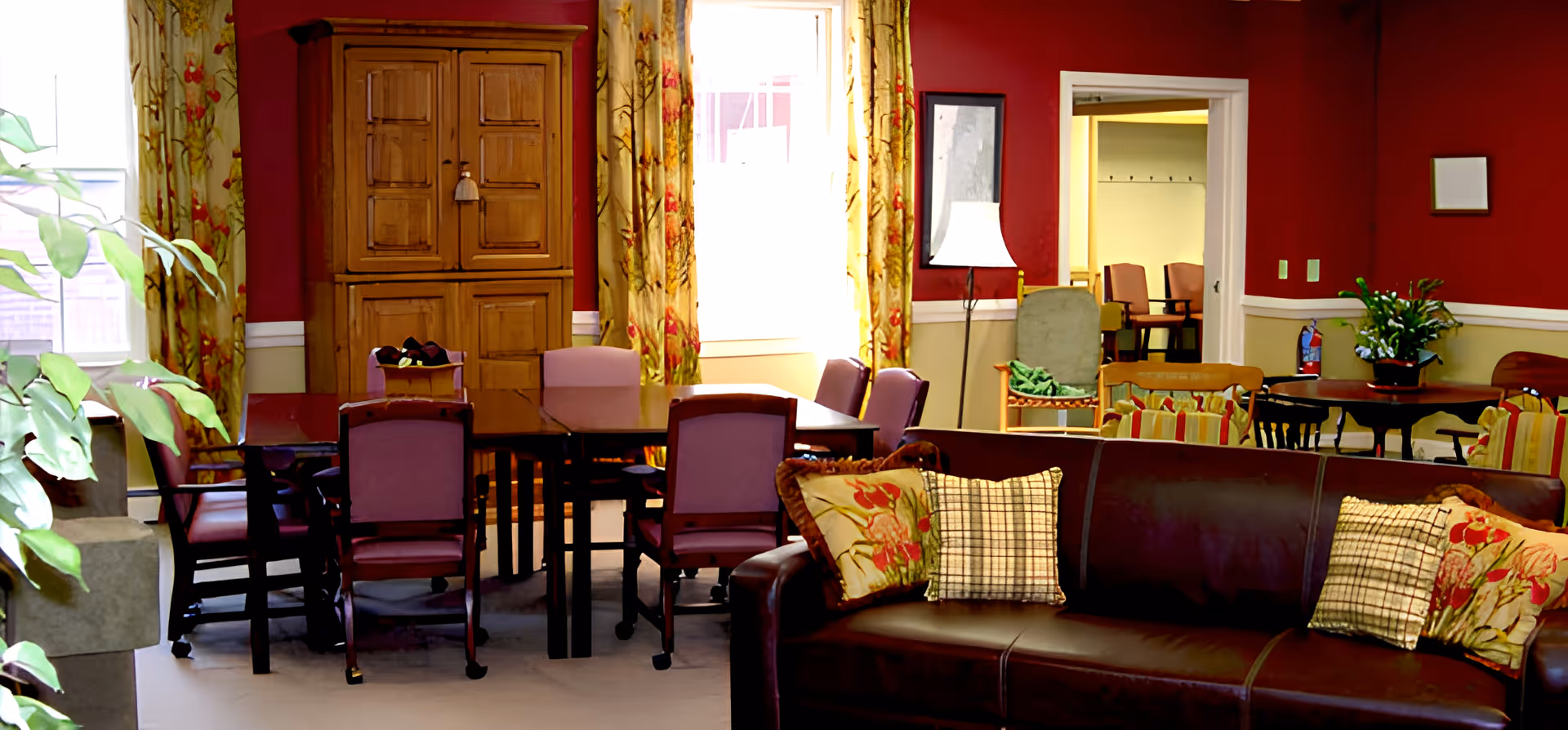 A cozy living room area with a dark brown leather sofa adorned with patterned pillows in the foreground. Behind the sofa is a dining table surrounded by purple cushioned chairs. The walls are painted red with floral curtains framing two large windows. A wooden cabinet stands against the wall, and a floor lamp is positioned near an open doorway leading to another room with additional chairs. There are plants and other seating arrangements visible in the background.