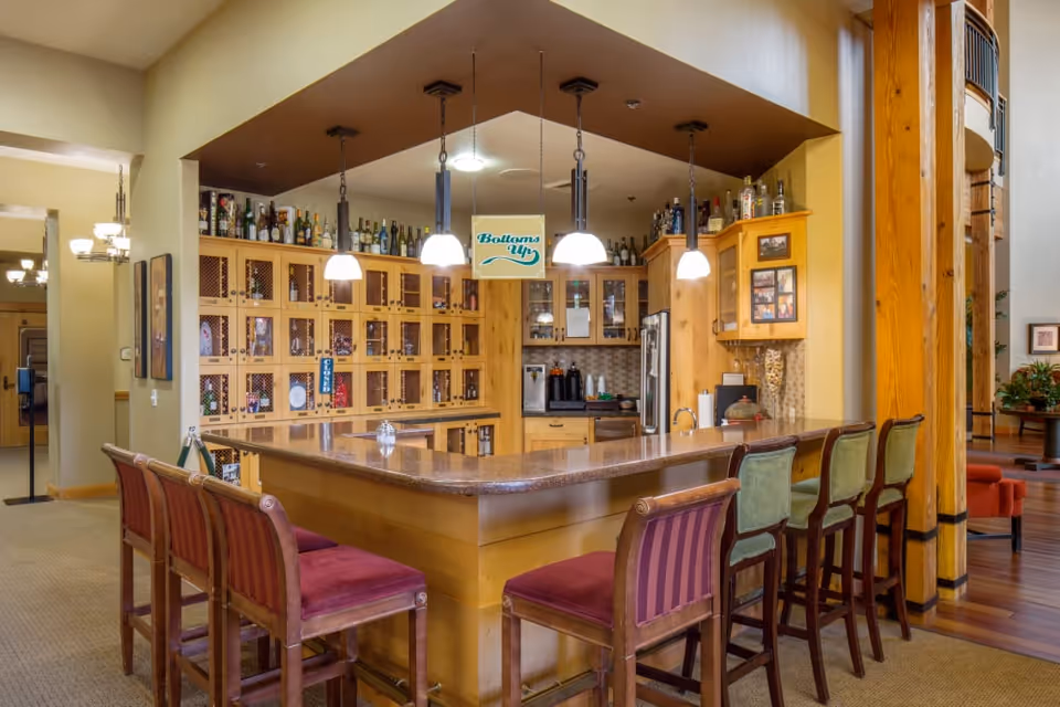 Interior view of a bar area in a senior living facility with a wooden counter surrounded by high chairs with red and green cushions. Behind the counter are wooden cabinets filled with bottles and glassware, and a sign hanging from the ceiling reads 'Bottoms Up'. The space is warmly lit with hanging pendant lights and has a cozy, inviting atmosphere.