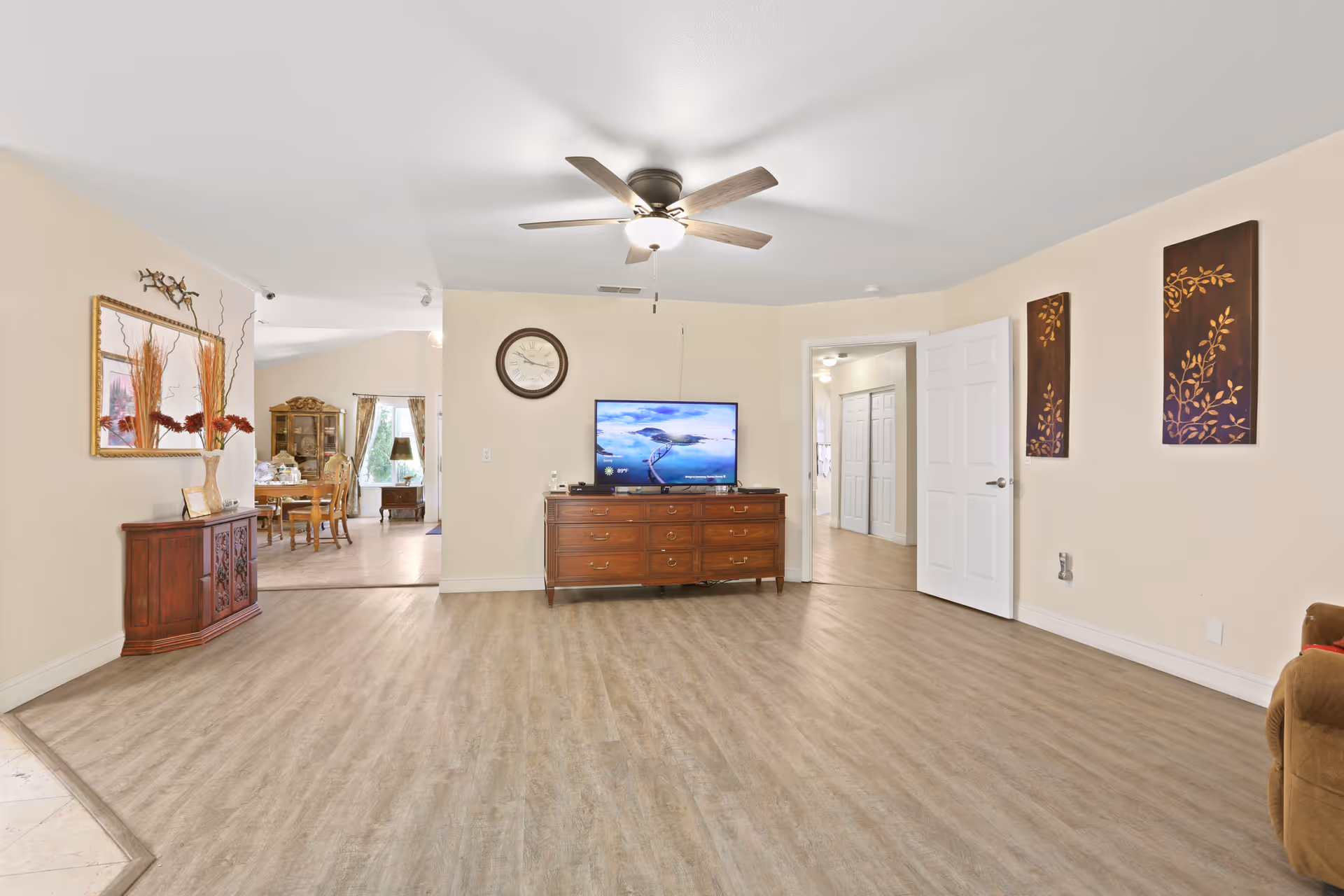 Spacious living room with wood-look flooring, ceiling fan, a dresser with a TV, and an open view into a dining area.
