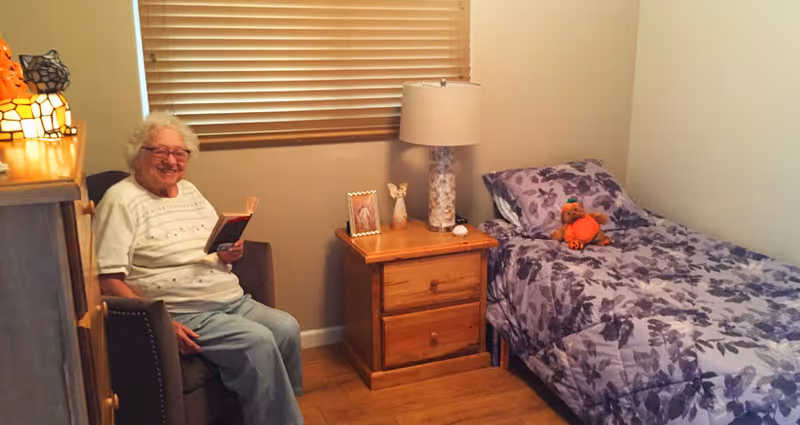 An elderly woman sitting in a chair reading a book in a cozy bedroom. The room has a single bed with purple floral bedding and a stuffed animal on it, a wooden nightstand with a lamp, a framed photo, and a small decorative item. The window has closed blinds.