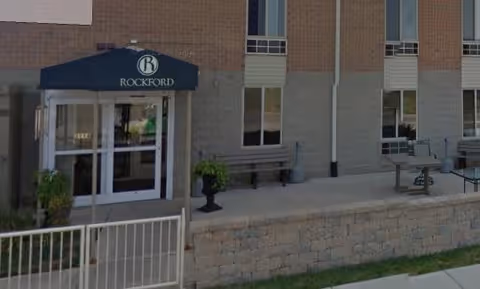 Front entrance of a brick building with a blue awning reading 'Rockford', a small patio with benches and planters.