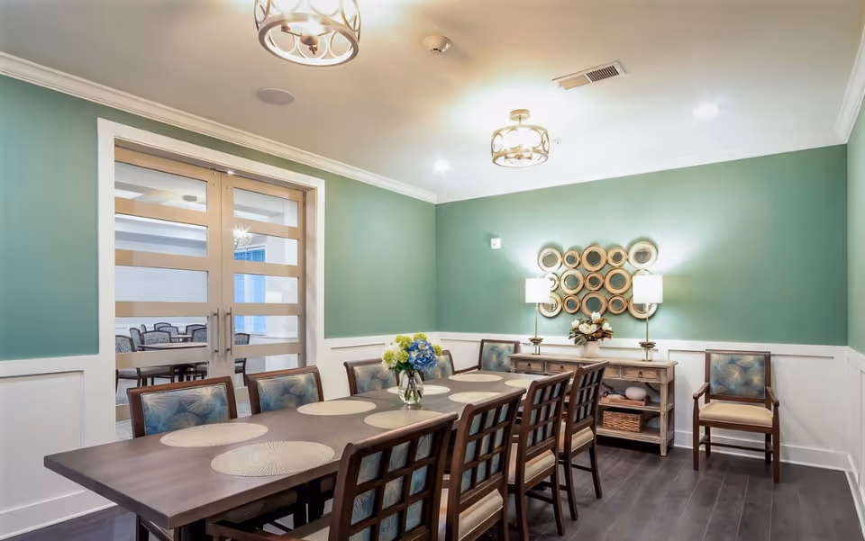 A dining room with a long wooden table surrounded by eight chairs with blue patterned cushions. The table has round placemats and a vase with flowers in the center. The walls are painted green with white wainscoting, and there are two modern ceiling light fixtures. A sideboard with two lamps and decorative items is against the far wall, which also features a circular mirror art piece. Glass double doors lead to another room with additional seating.