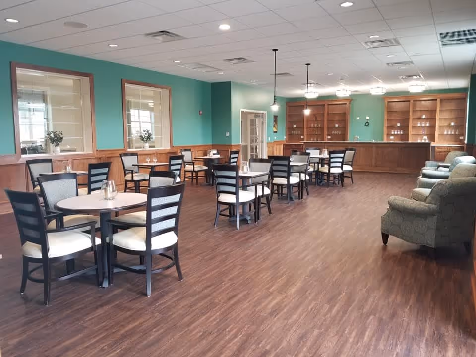 A spacious dining room with several round tables and chairs arranged neatly on a wooden floor. The walls are painted green with wood paneling on the lower half. There are large windows on one side with plants on the windowsills. At the far end, there is a wooden counter with shelves behind it, and three pendant lights hanging from the ceiling. On the right side, there are a few upholstered armchairs.