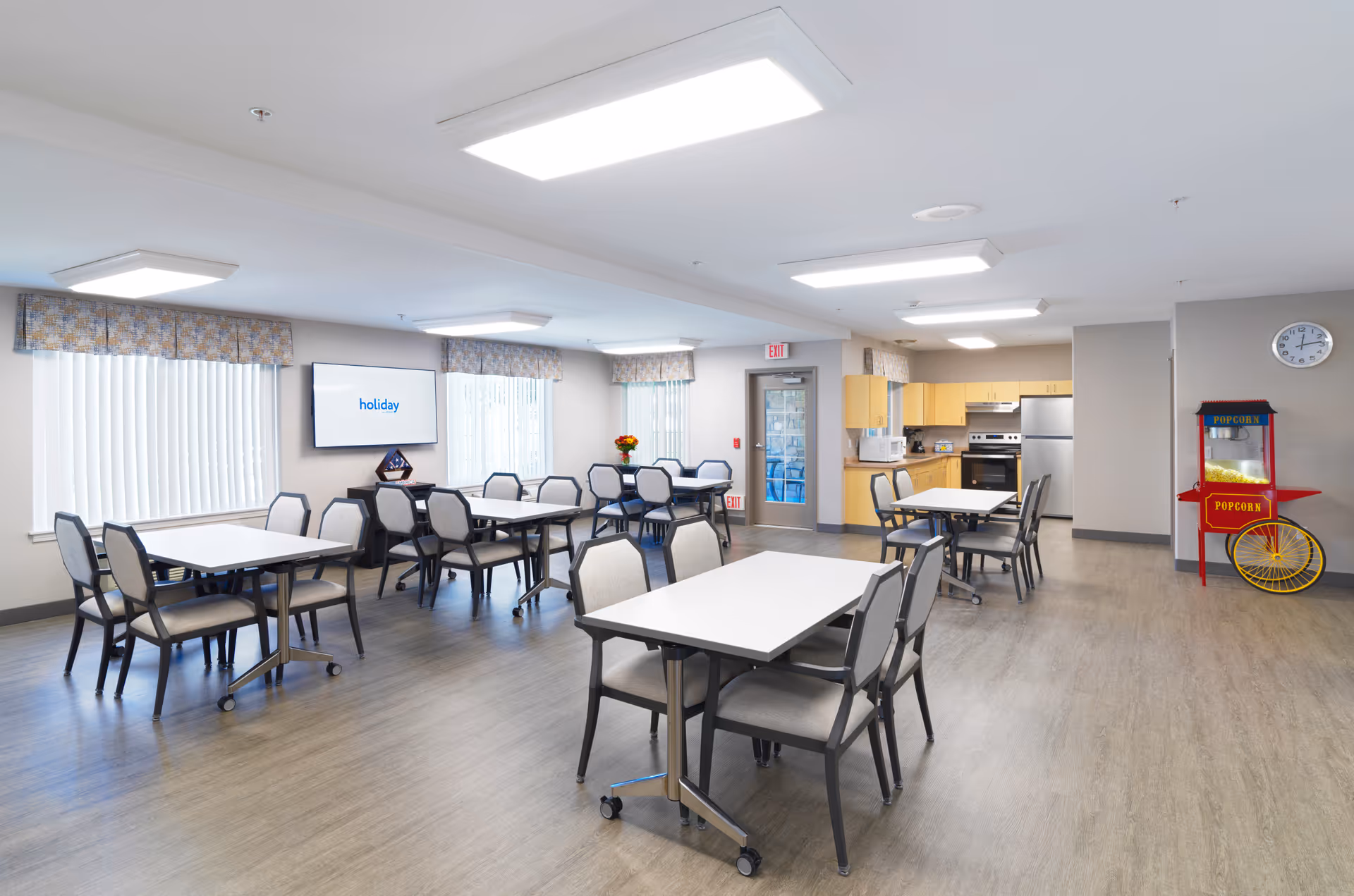A bright and spacious dining area in a senior living facility with several tables and chairs arranged neatly. The room features large windows with vertical blinds and valances, a wall-mounted TV displaying the word 'holiday', a popcorn machine in the corner, and a kitchenette with light wood cabinets and stainless steel appliances.