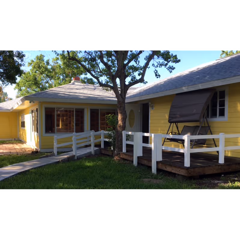 Exterior view of a yellow single-story building with a gray roof, white railings, a wooden deck with a covered swing, and a tree in the foreground. The building is surrounded by green grass and trees under a clear blue sky.