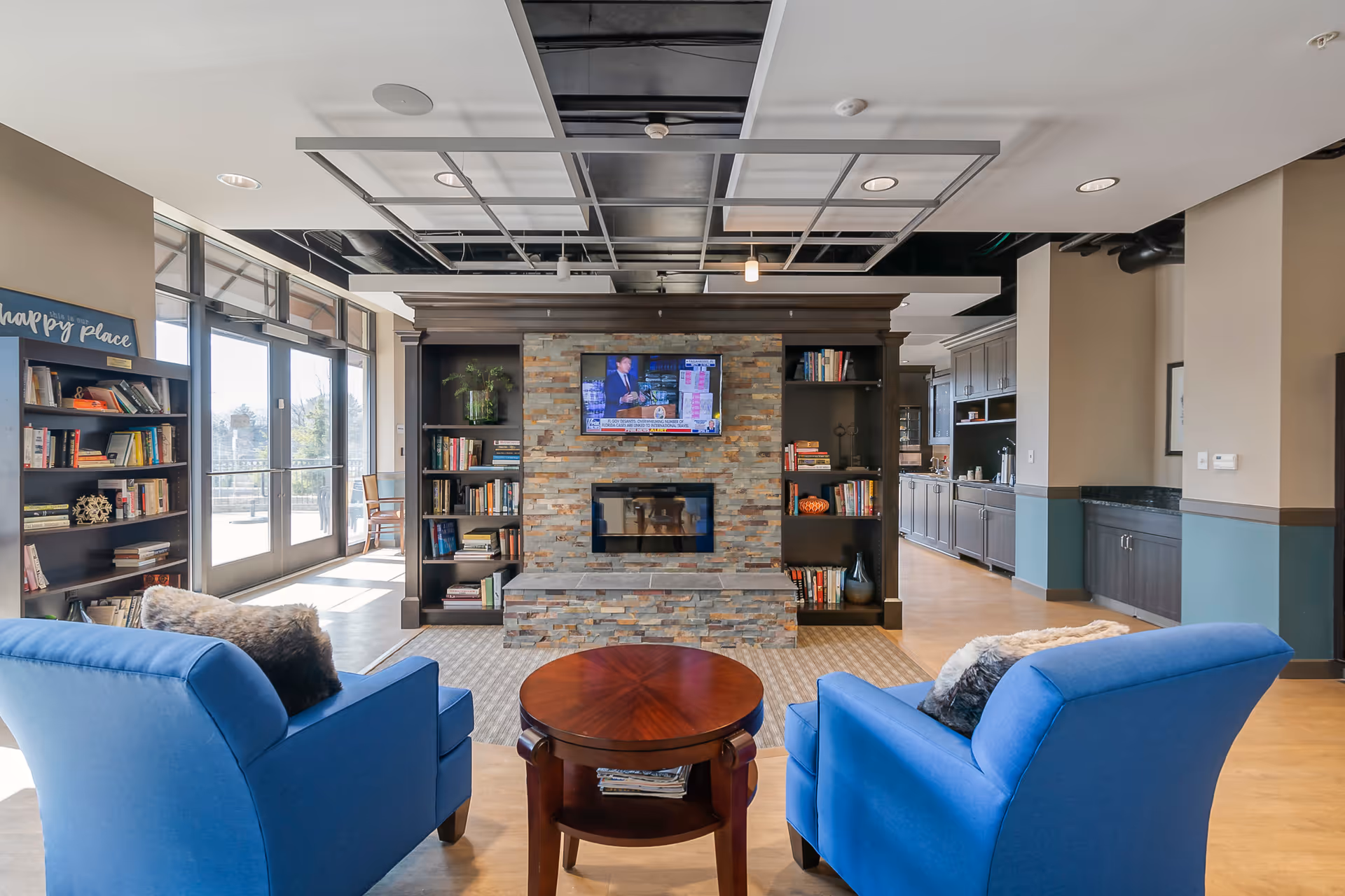 A cozy living room area with two blue armchairs facing a stone fireplace with a mounted TV above it. The fireplace is flanked by built-in dark wood bookshelves filled with books and decorative items. A round wooden coffee table is placed between the chairs. Large windows and glass doors on the left side let in natural light, and a kitchen area is visible in the background on the right.