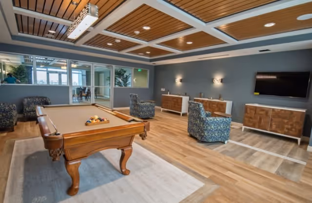 A spacious recreational room with a wooden pool table in the center, surrounded by patterned armchairs. The room features a wooden ceiling with recessed lighting, a large flat-screen TV mounted on the wall, and modern wooden cabinets. The floor is a combination of wood and area rugs, and large windows allow natural light to enter the space.