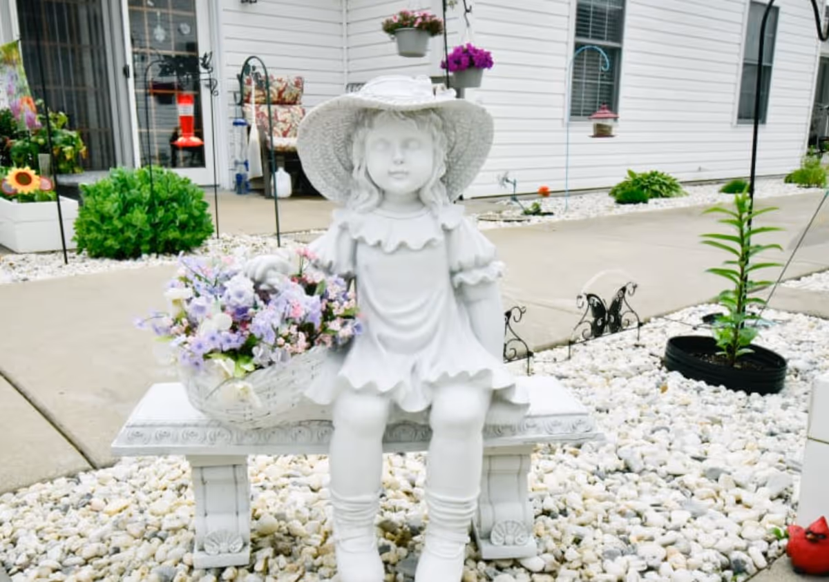 White statue of a girl wearing a hat sits on a bench in a front garden.