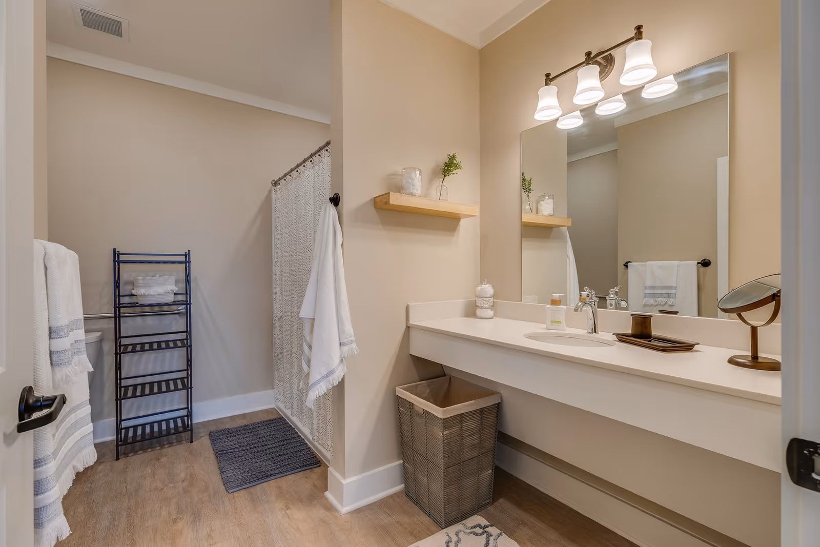 Well-lit bathroom with a single-sink vanity and large mirror, shower with curtain, towel racks, shelving, and a wicker waste basket.