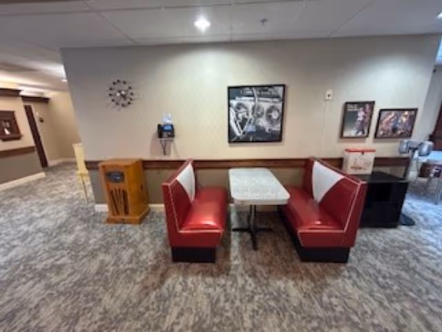 Interior view of a seating area in a senior living facility featuring two red and white cushioned booths facing each other with a rectangular table in between. The area has carpeted flooring and beige walls adorned with framed pictures and a clock. There is a wooden cabinet and a wall-mounted telephone on the left side, and a water dispenser on the right side.