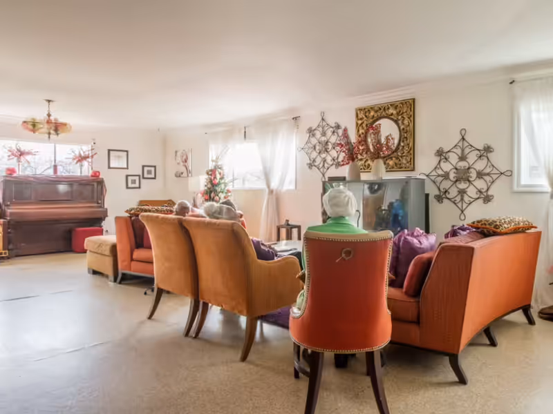 Cozy senior living room with orange upholstered chairs arranged around a television, a piano in the corner, and decorative wall accents.