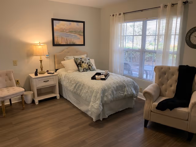 A cozy bedroom with a single bed covered in a light blue quilt and decorative pillows. A tray with a cup and a book rests on the bed. To the left of the bed is a white nightstand with a lamp, a small black cat figurine, and a phone. There is a beige tufted chair next to the nightstand and a larger beige armchair with a black throw on the right side near a large window with sheer white curtains. A framed painting of a beach scene hangs above the bed. The floor is wood, and the room is softly lit.