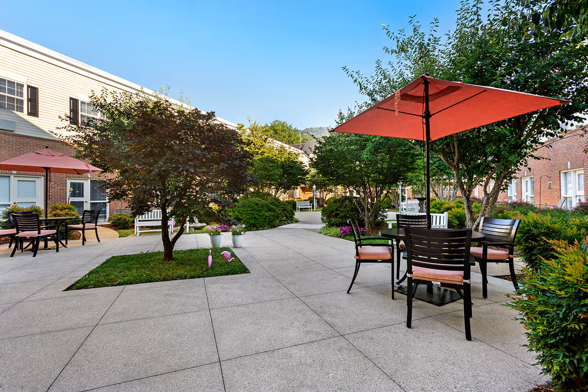 Outdoor courtyard area at StoryPoint Chattanooga featuring paved walkways, green bushes, trees, and several seating areas with tables and chairs under red umbrellas. The courtyard is surrounded by brick and siding buildings under a clear blue sky.