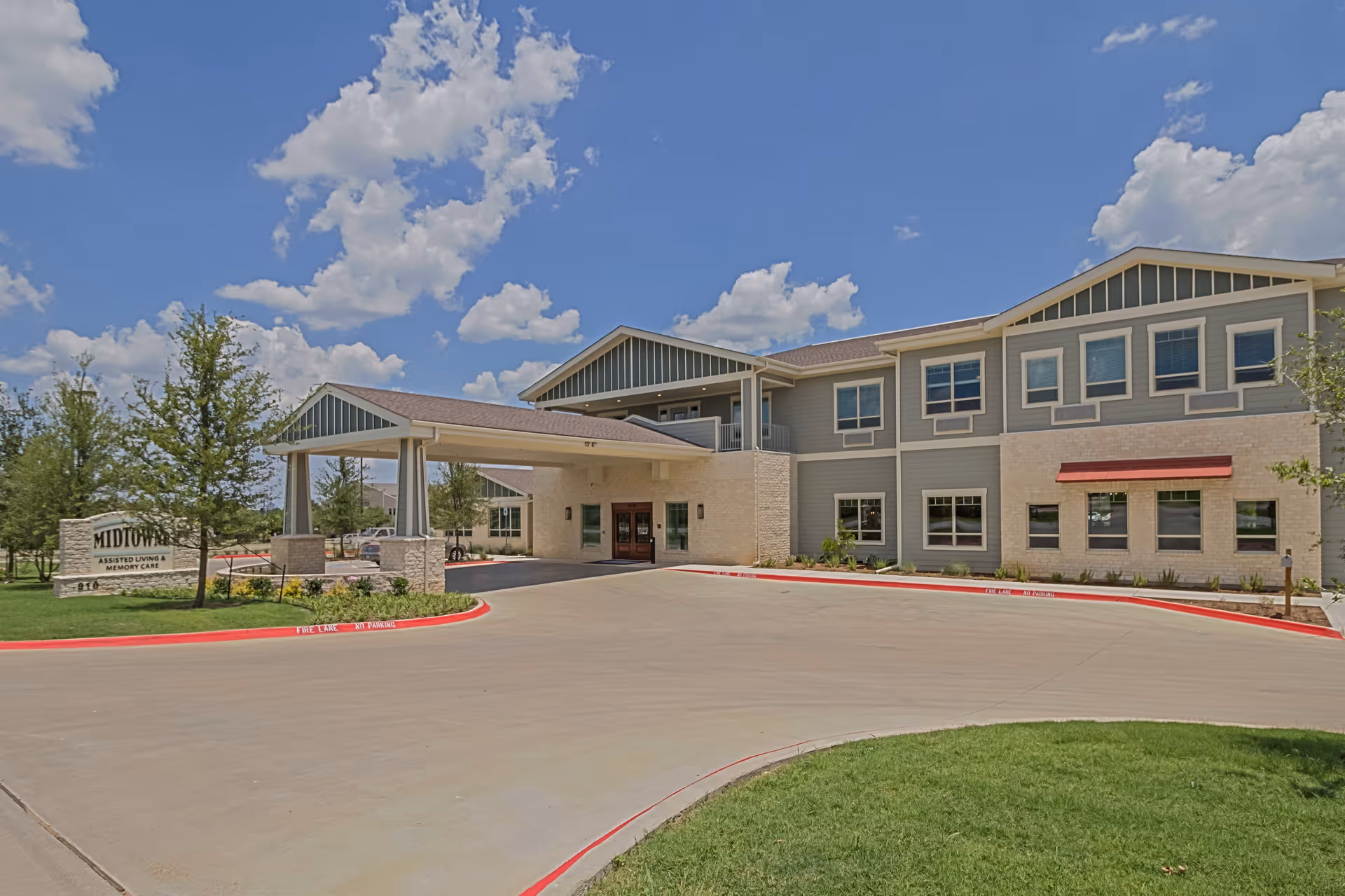 Exterior view of Midtowne Assisted Living and Memory Care facility showing a two-story building with a covered entrance, surrounded by a driveway, green lawn, and trees under a partly cloudy blue sky.