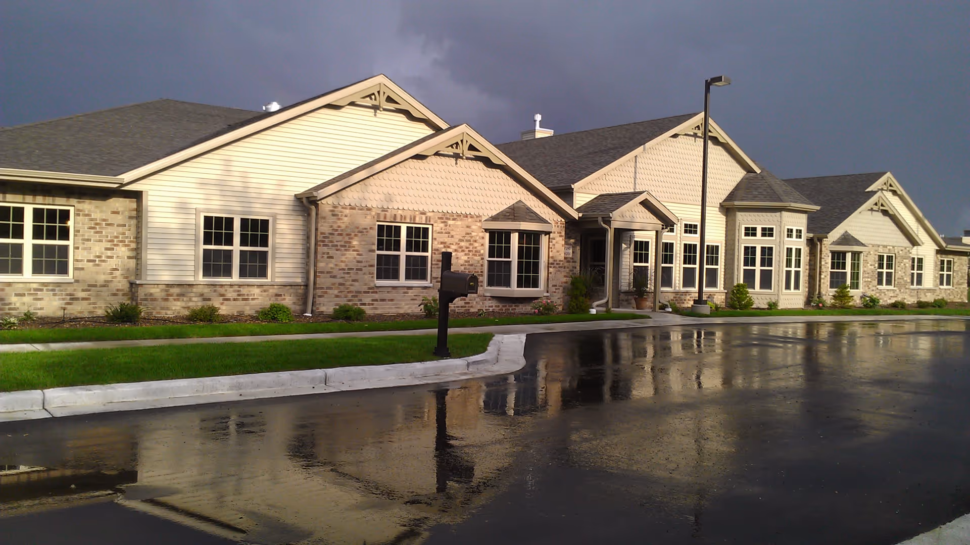 Exterior view of a single-story assisted living facility building with beige siding and brick accents, multiple windows, a mailbox, and a wet driveway reflecting the building under a cloudy sky.