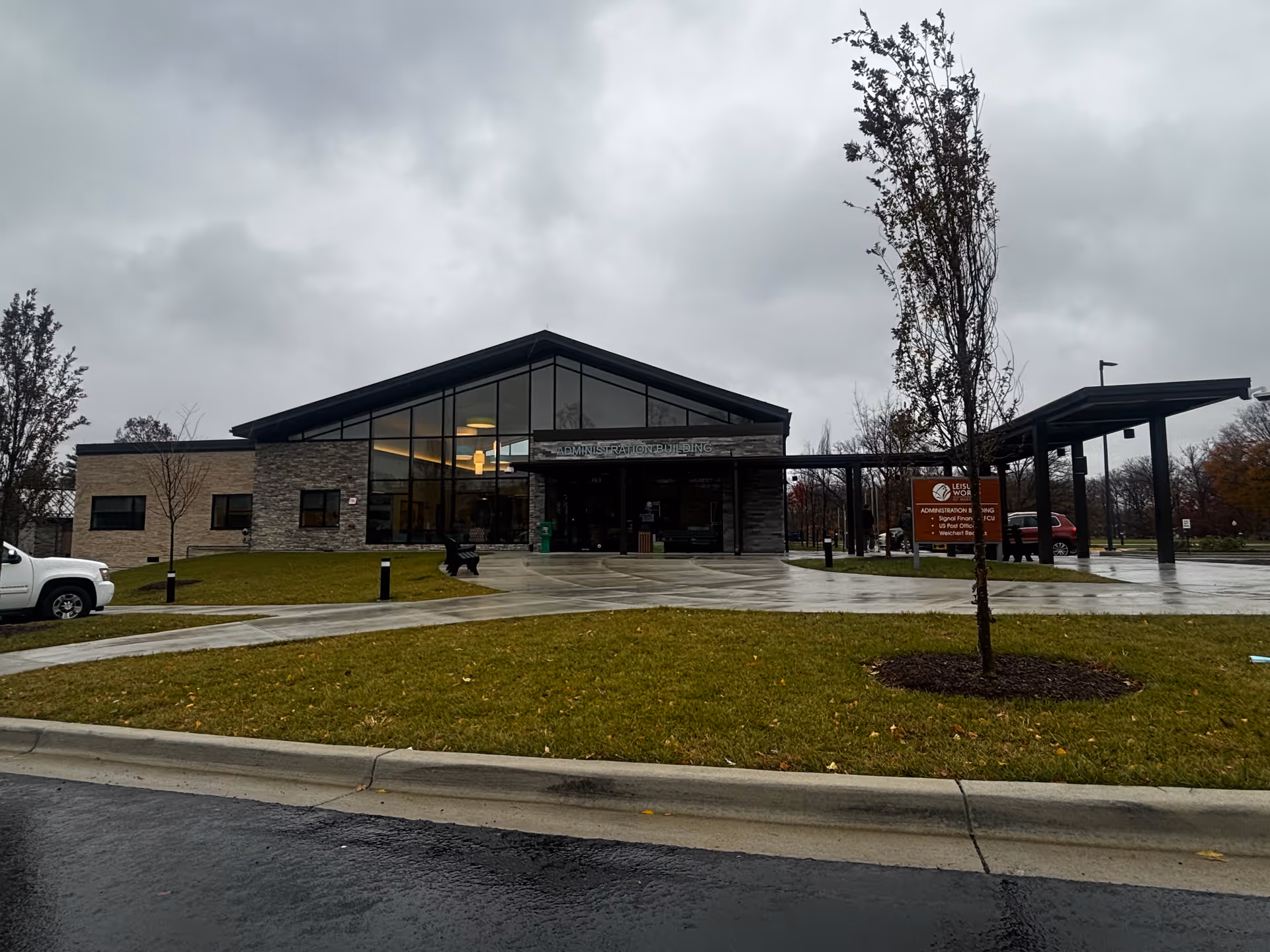Exterior view of the Leisure World of Maryland administration building on a cloudy day. The building has large glass windows, stone and brick walls, and a covered entrance. There is a green lawn with young trees and a paved driveway in front. A white vehicle is partially visible on the left side.