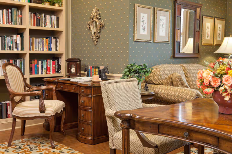 A cozy and elegant interior room featuring a wooden desk with papers and decorative items, a striped upholstered chair, a bookshelf filled with books, a patterned sofa, framed floral artwork on the wall, a mirror, a table with a floral arrangement, and a green wallpaper with a subtle gold pattern.