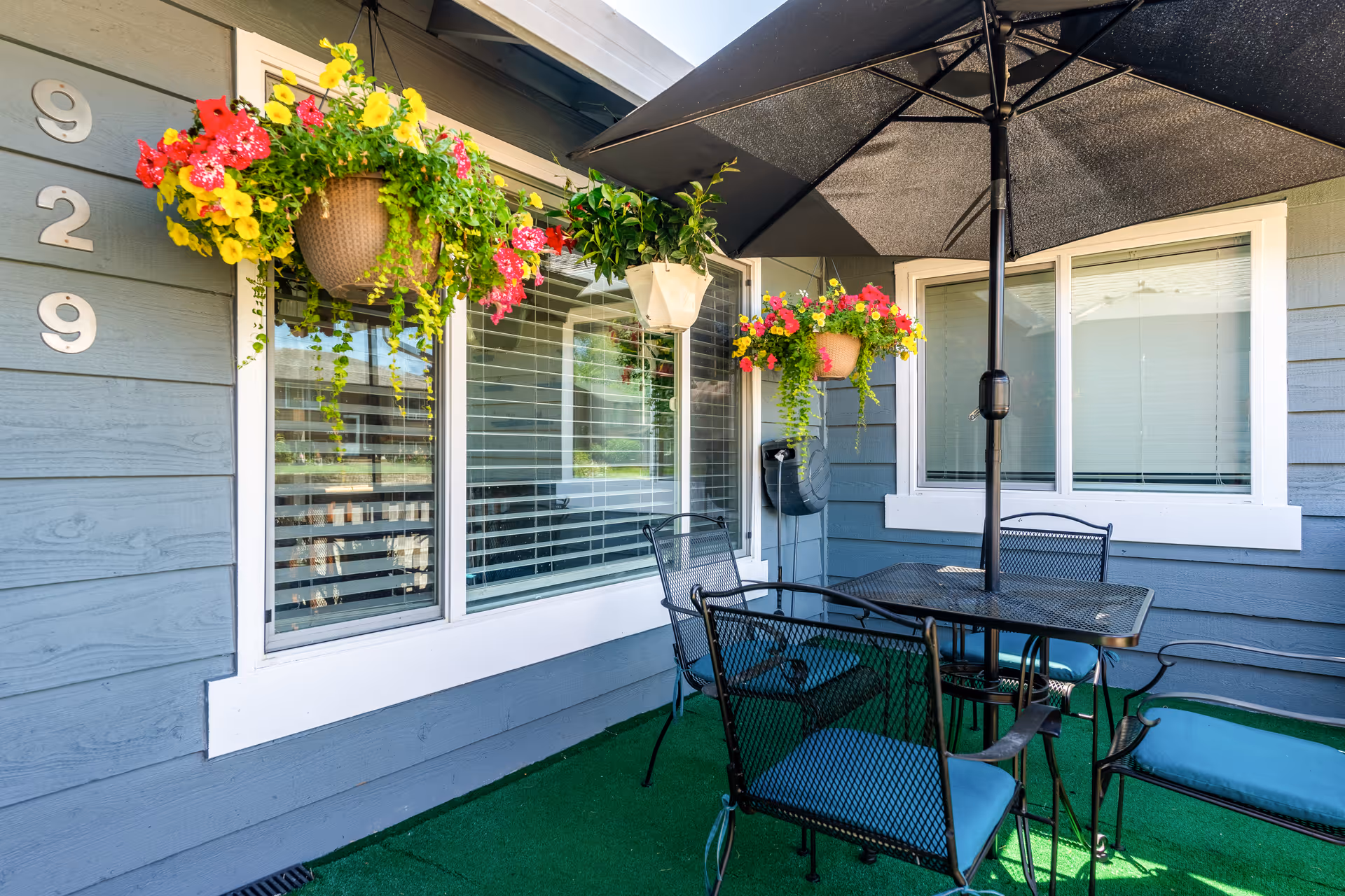 Small outdoor patio with a black umbrella over a metal table and chairs, hanging flower baskets, and gray siding marked 929.