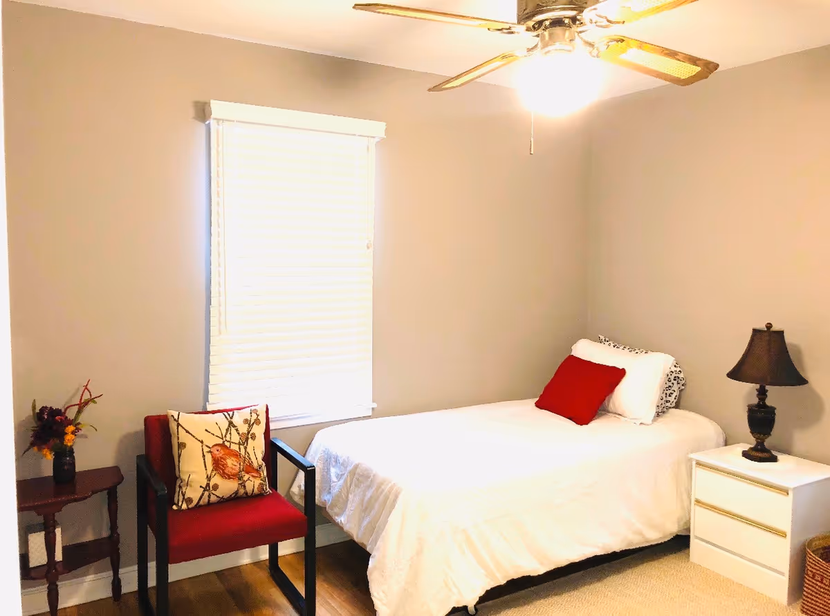 A small bedroom with a single bed covered in white bedding and two pillows, one white and one red. Next to the bed is a white nightstand with a dark lamp on it. A red chair with a decorative pillow featuring a bird design is placed near a window with closed blinds. There is a small wooden side table with a vase of flowers beside the chair. The room has beige walls, wooden flooring, and a ceiling fan with a light.