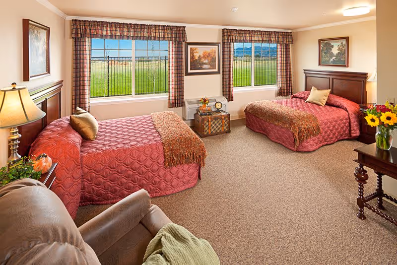 A cozy bedroom in a memory care community featuring two twin beds with red quilted bedspreads and decorative pillows. The room has two large windows with plaid curtains, allowing natural light to brighten the space. Between the beds is a small chest with a clock and a flower arrangement. There is a comfortable armchair with a green throw blanket in the foreground, a side table with a lamp and a vase of sunflowers, and framed artwork on the walls. The carpeted floor and warm lighting create a welcoming atmosphere.