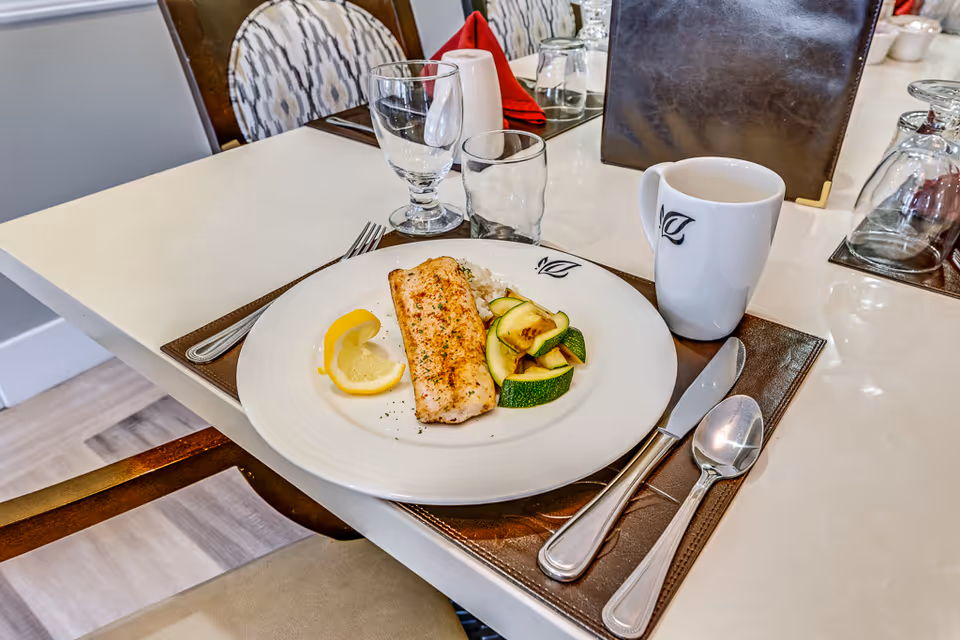 A dining table set with a plate of grilled fish, lemon wedges, grilled zucchini, and rice. The table also has a white coffee mug, a glass, a water glass, a fork, a knife, and a spoon on a brown placemat. Chairs with patterned upholstery are visible in the background.