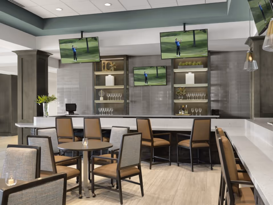 Modern lounge area with a bar counter and multiple chairs around small round tables. Three flat-screen TVs mounted on the wall display a golfer in action. Shelves behind the bar hold decorative items, wine bottles, and glasses. The space features neutral tones with gray tiled walls and light wood flooring.
