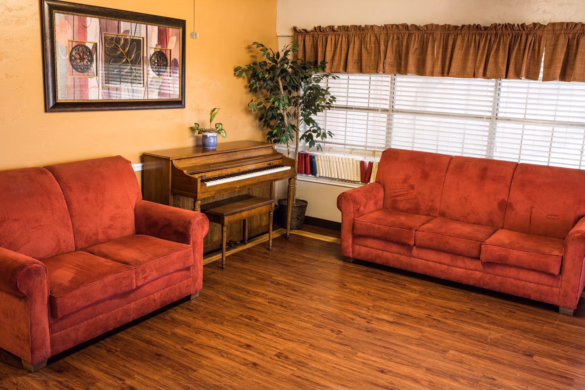 A cozy living room with two red upholstered sofas facing each other, a wooden piano with a matching bench, a potted plant, a large window with blinds and brown valance, and a framed abstract artwork on a yellow wall.