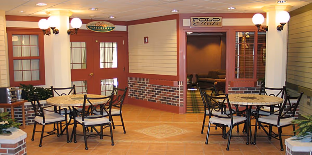 Interior common area with round tables and chairs on a tiled floor in front of doorways and signs reading "Waterford" and "Polo Club".