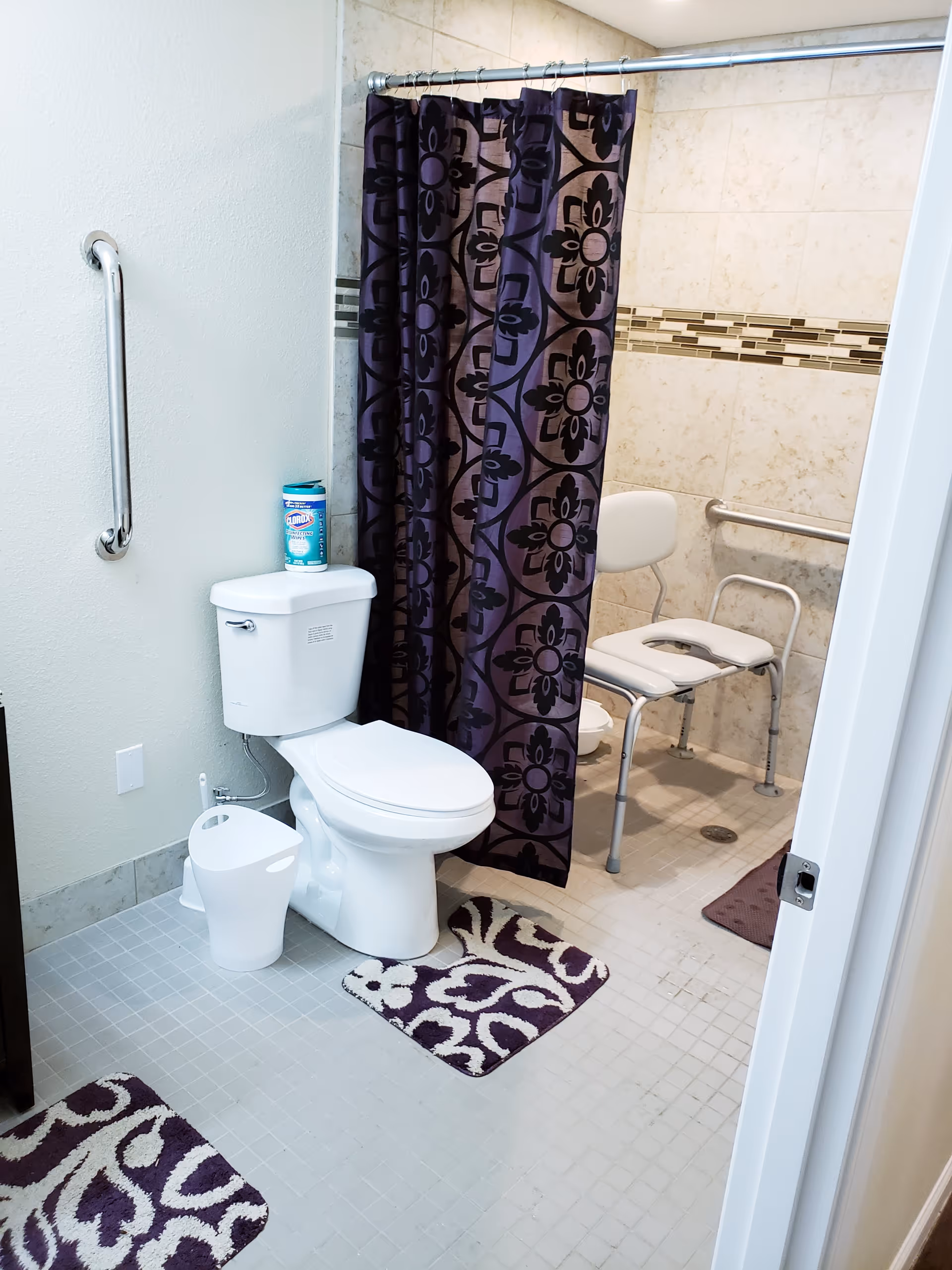 A bathroom with a white toilet, a purple and black patterned shower curtain, and a white shower chair inside a tiled shower area. There are two purple and white patterned bath mats on the floor, a grab bar on the wall next to the toilet, and a container of disinfecting wipes on top of the toilet tank.