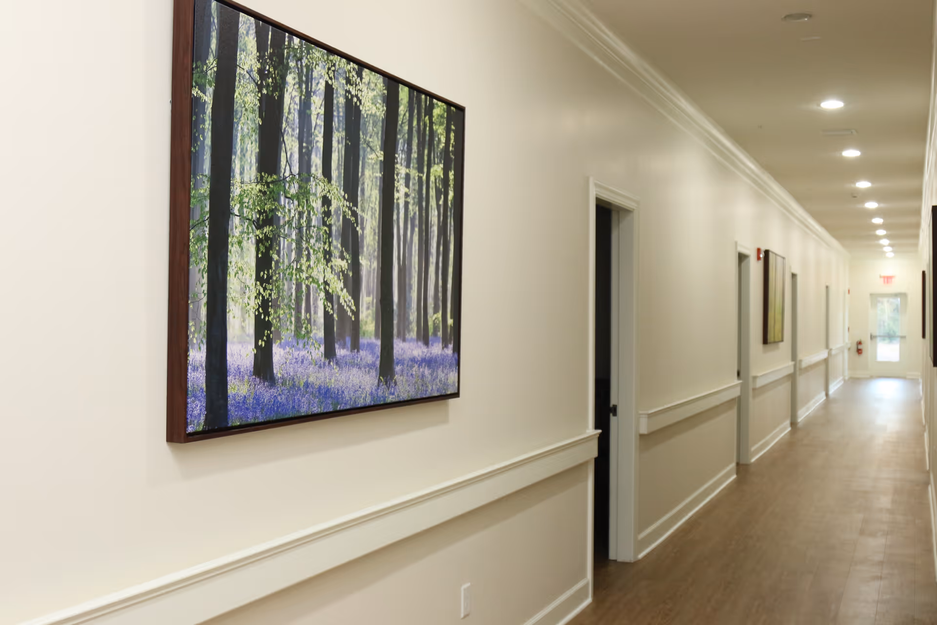 A long, well-lit hallway in a senior living facility with light-colored walls and wooden flooring. The hallway features several open doorways and framed nature artwork on the walls. Recessed ceiling lights illuminate the corridor, and an exit door is visible at the far end.