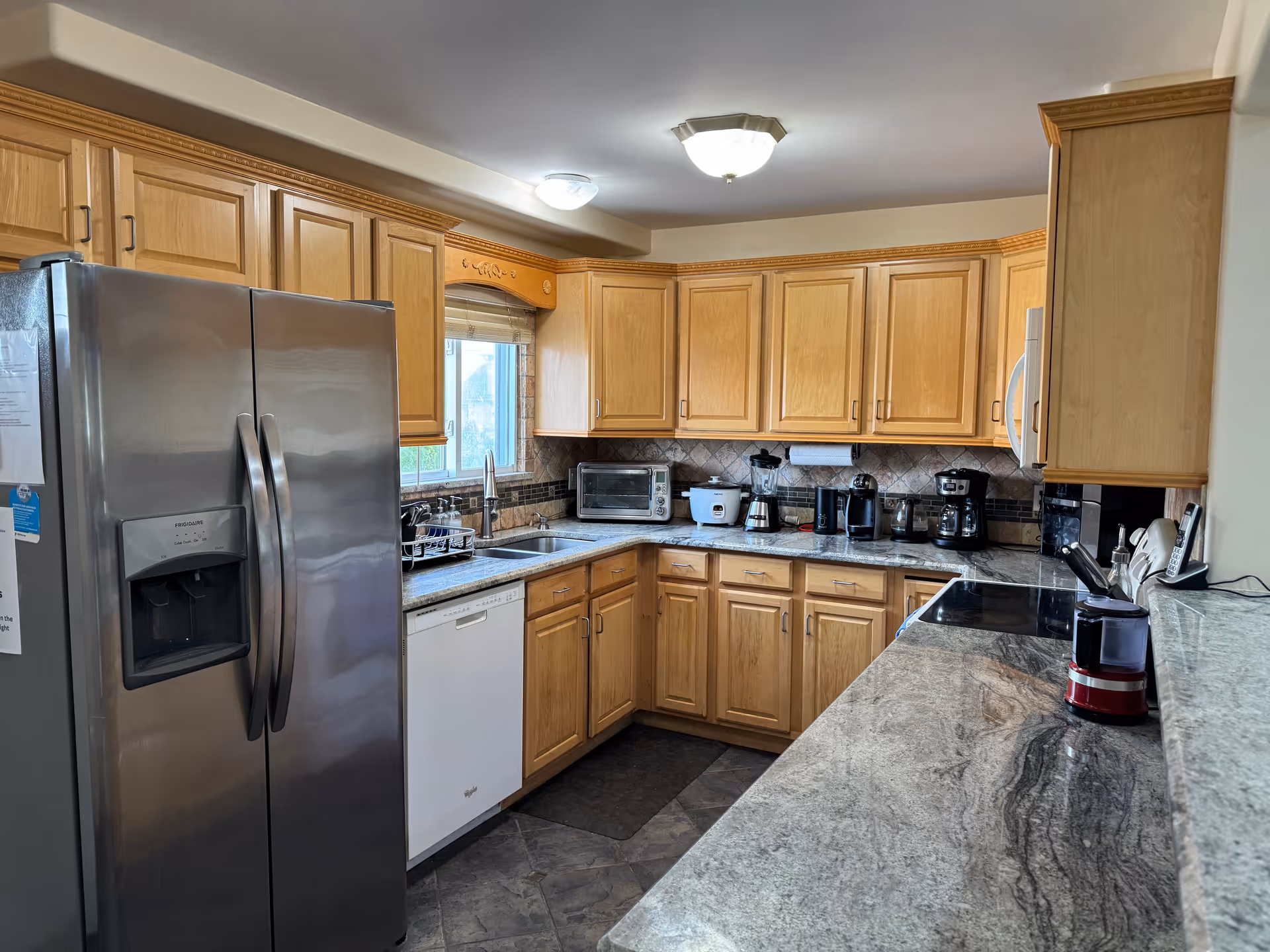 A modern kitchen with light wood cabinets, a stainless steel refrigerator with water and ice dispenser, a white dishwasher, a double sink under a window, and various small kitchen appliances on the granite countertops including a toaster oven, rice cooker, blender, coffee makers, and a red coffee grinder.