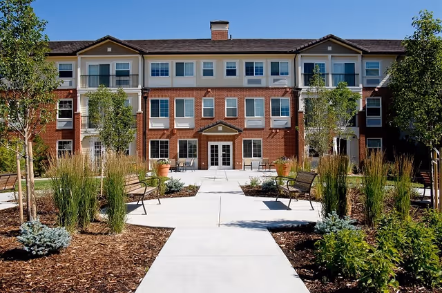 Front exterior view of a three-story senior living facility building with a brick and beige facade, multiple windows, a central entrance with double doors, and a landscaped walkway with benches and plants leading up to the entrance.