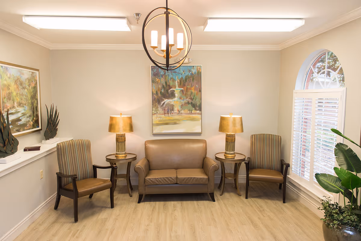 A small seating area with a leather loveseat flanked by two striped armchairs, side tables with lamps, framed artwork, and a window with shutters.