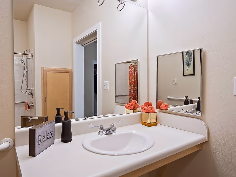 Bathroom sink with a faucet, soap dispenser, and a decorative block with the word 'Relax'. A mirror above the sink reflects part of the bathroom, including a shower with a red patterned shower curtain and a towel hanging on the shower rod. The walls are light-colored, and there is a small framed picture visible in the reflection.