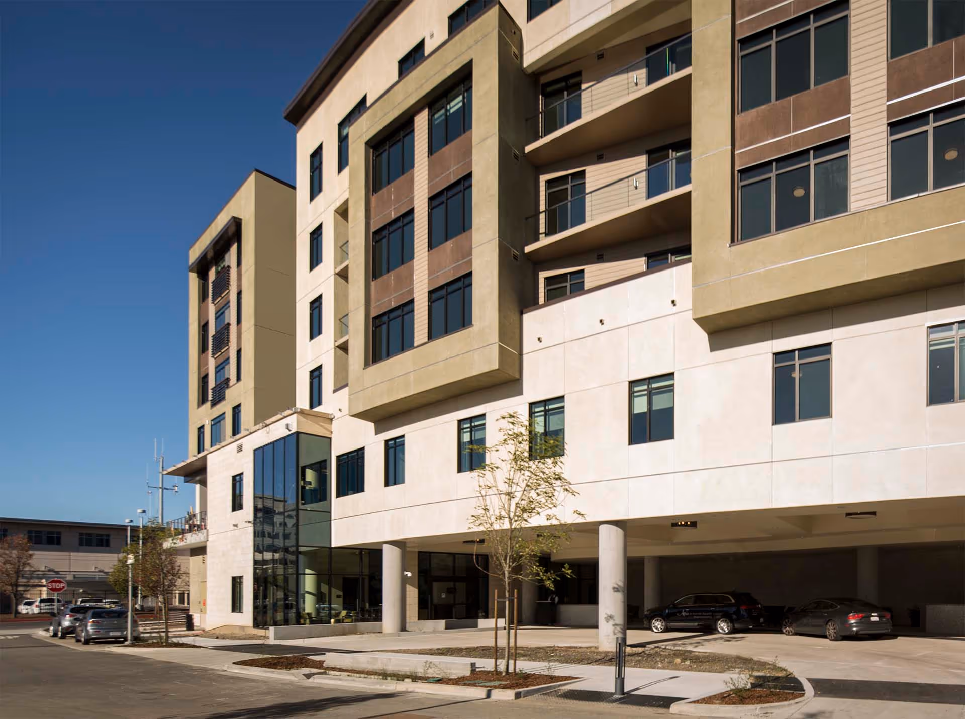 Exterior view of a modern multi-story building with large windows and balconies, featuring a covered parking area underneath. There are a few cars parked and small trees planted along the sidewalk under a clear blue sky.