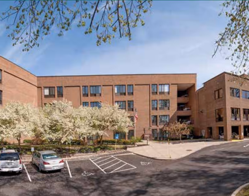 Front view of a multi-story brick senior living building with flowering trees, parking spaces, and an American flag.