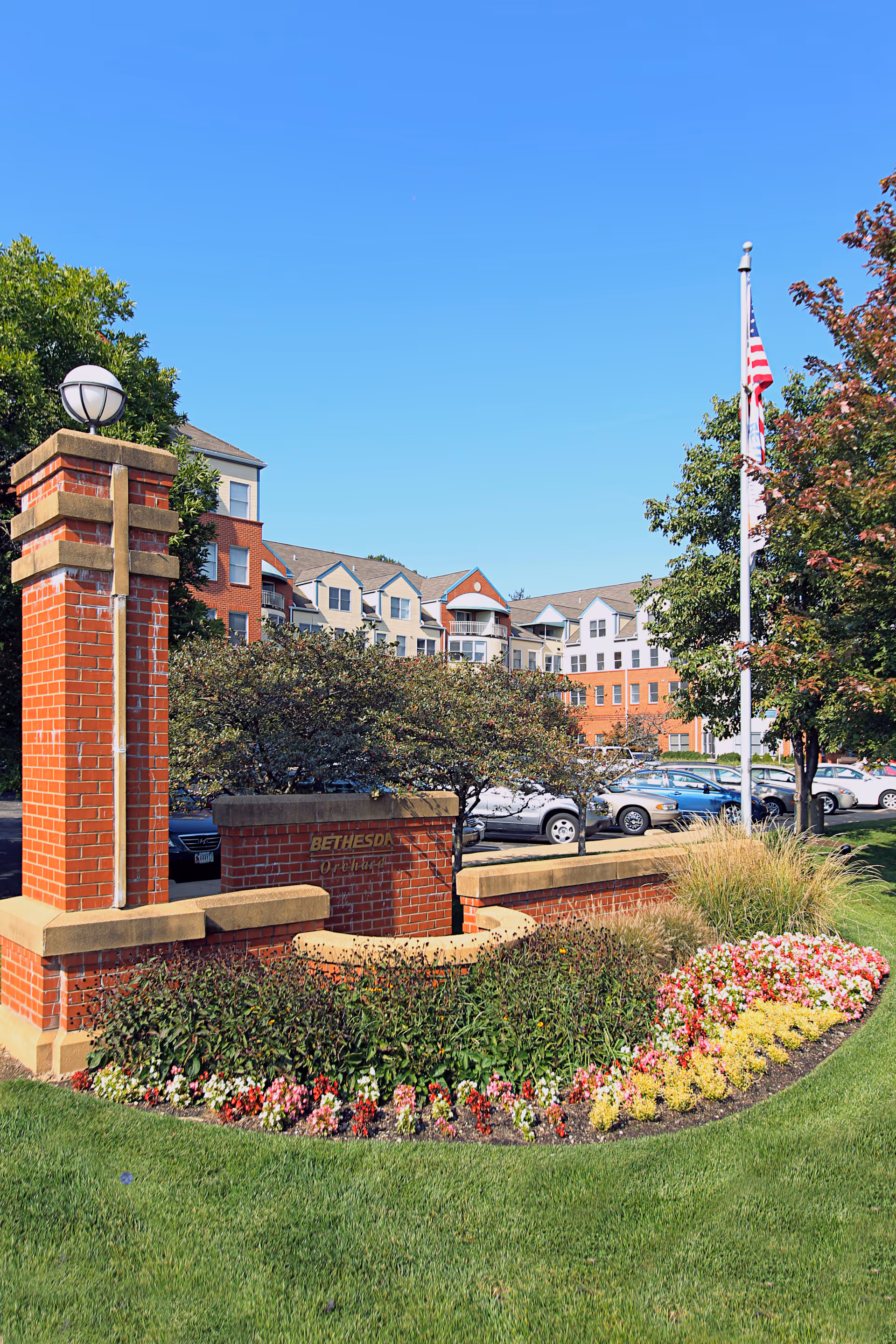 Brick entrance sign and landscaped flowerbed in front of a multi-story senior living building with parked cars and an American flag.