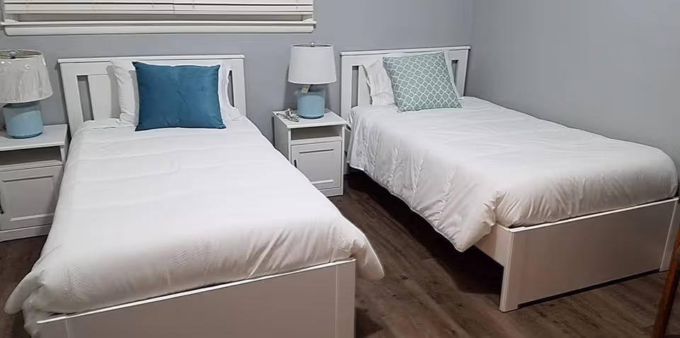 A bedroom with two single beds, each with white bedding and a decorative pillow, one blue and one patterned. Between the beds is a white nightstand with a lamp and some items on it. The room has wooden flooring and a window with closed blinds above the beds.