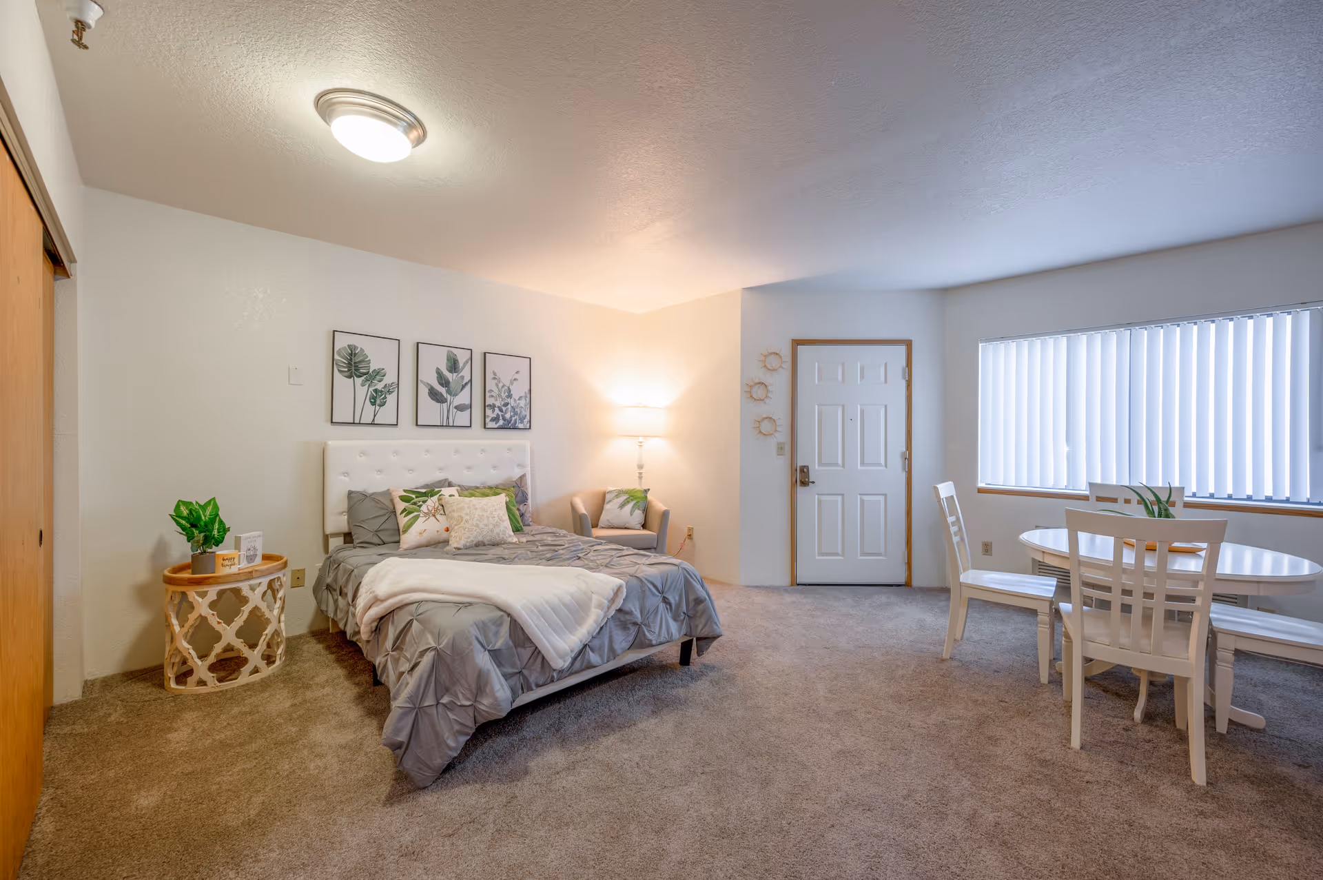 A spacious studio apartment with a bed featuring a tufted white headboard, gray bedding, and decorative pillows. Next to the bed is a round side table with a plant and a small framed picture. On the wall above the bed are three framed botanical prints. A cozy armchair with a pillow is placed in the corner near a floor lamp. To the right, there is a white round dining table with four chairs near a large window with vertical blinds. The entrance door is visible in the background. The room has beige carpet and white walls.