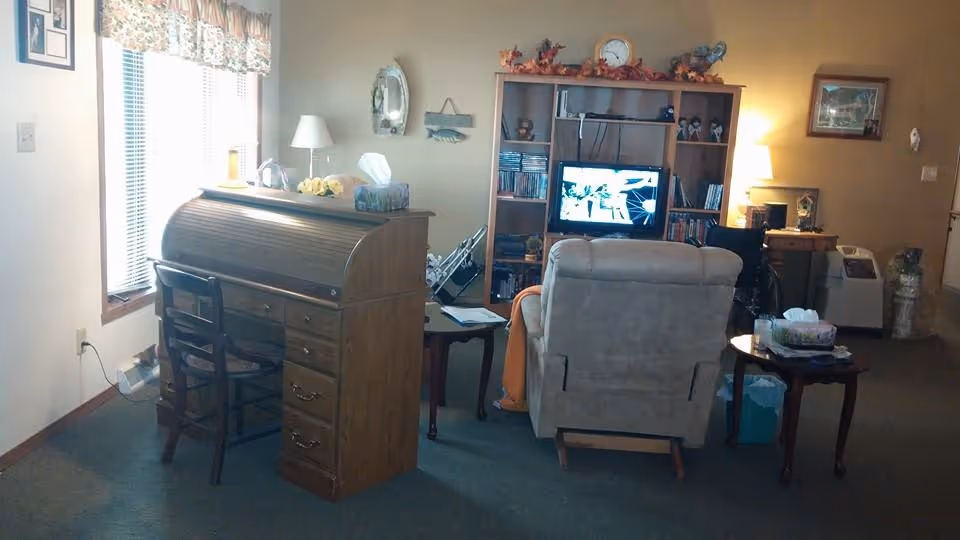 A cozy living room area with a wooden roll-top desk and chair near a window with floral curtains. In the background, there is a beige recliner facing a wooden entertainment center with a TV and various decorative items. A side table with a lamp and tissues is next to the recliner, and another lamp is on a desk in the corner. The room has carpeted flooring and warm lighting.