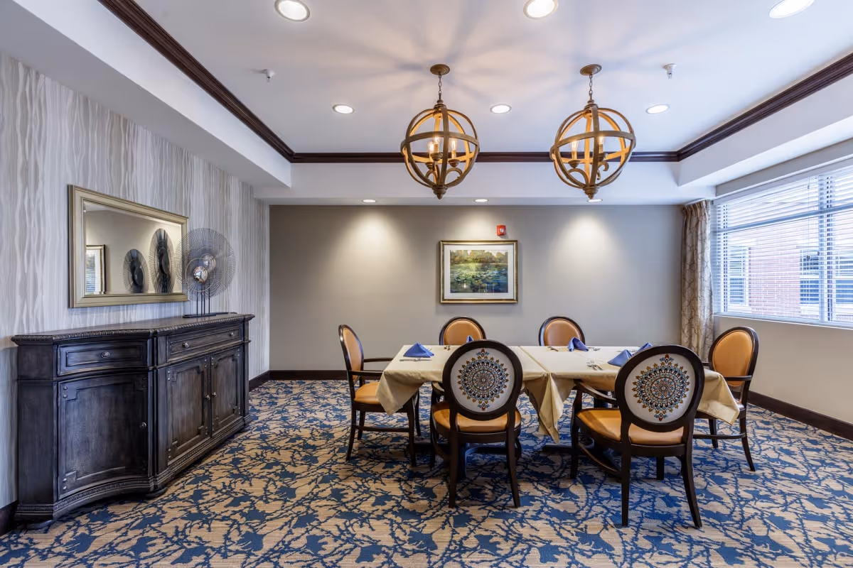 A dining room with a beige-clothed table surrounded by decorative chairs, pendant lights overhead, a sideboard and a large window.