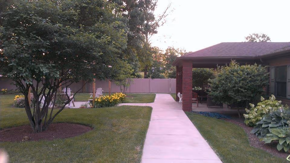 Outdoor area of a senior living facility featuring a concrete walkway leading to a covered patio with seating. The area is landscaped with green grass, trees, bushes, and flower beds. A fence is visible in the background.