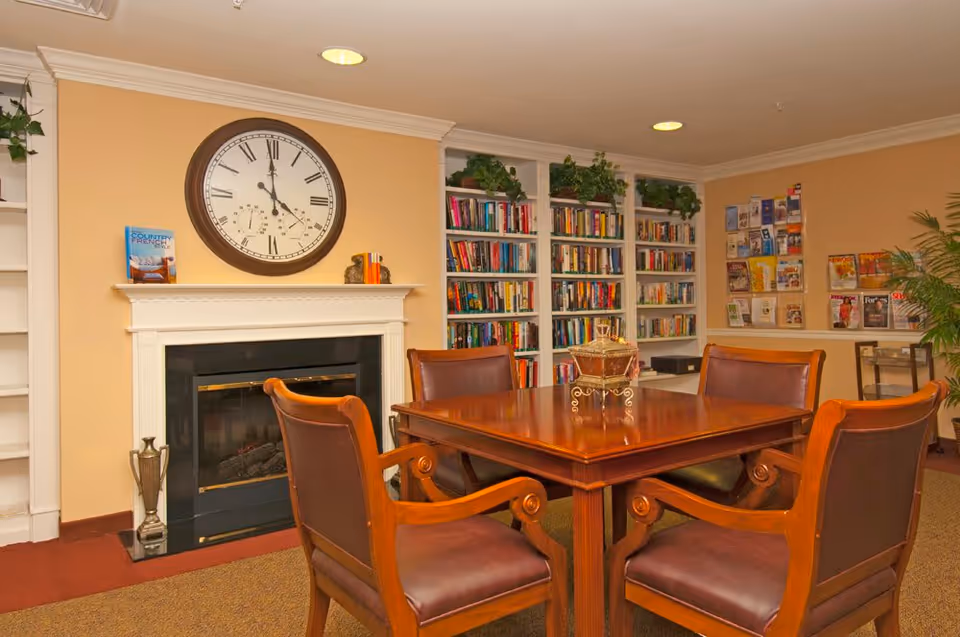 A cozy interior room with a wooden table and four leather chairs arranged around it. Behind the table is a fireplace with a large round clock above it. To the right of the fireplace are built-in bookshelves filled with books and some green plants on top. On the far right wall, there are racks with magazines and brochures. The walls are painted a warm beige color and the room is well-lit with recessed ceiling lights.