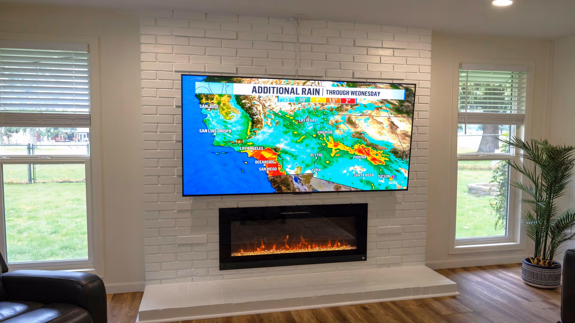 A modern living room area with a white brick wall featuring a mounted flat-screen TV displaying a weather map. Below the TV is a sleek electric fireplace with visible flames. The room has two tall windows on either side of the fireplace, letting in natural light and showing a green outdoor lawn. There is a potted plant on the right side and part of a black leather chair visible on the left.