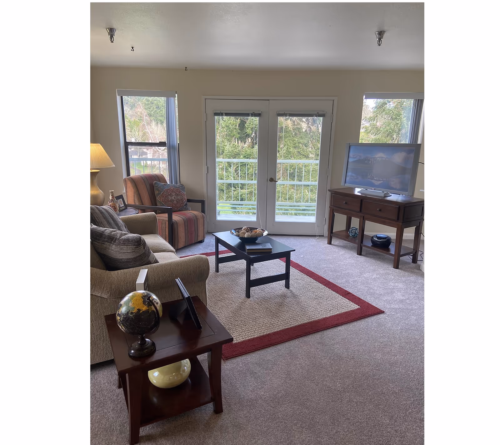 Cozy living room with a sofa, armchair, coffee table, TV on a wooden stand, and double glass doors opening to a balcony with trees beyond.